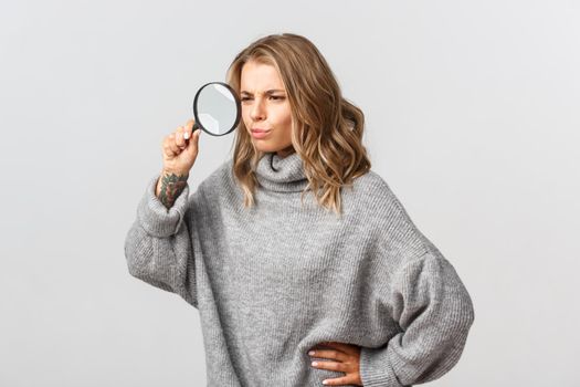 Attractive blond girl searching for something with magnifying glass, standing serious over white background.