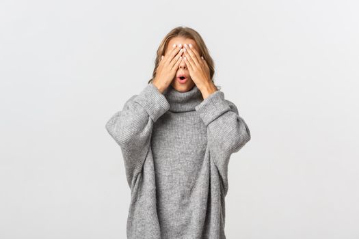 Portrait of beautiful young woman in grey sweater, standing blindsided with hands on face, waiting for surprise, standing over white background.