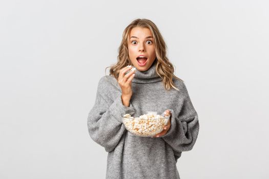 Image of amazed blond girl watching movie, looking excited, eating popcorn, standing over white background.