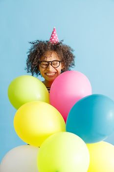 Joyful multiethnic lady in birthday cone hat keeping eyes closed and smiling while holding colorful balloons. Isolated on blue background