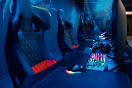 View on tables equipped with PC monitors, brightly lit keyboards and headsets with empty streaming chairs in dark gaming room