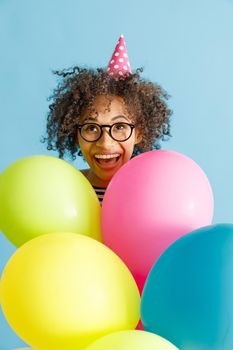 Cheerful multiethnic lady in birthday cone hat holding party balloons. Isolated on blue background