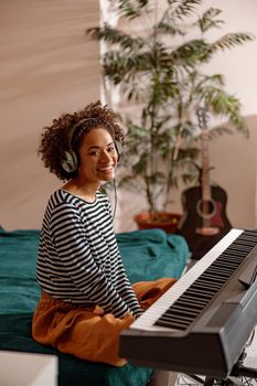 Cheerful multiracial female musician looking at camera and smiling while sitting on bed in front of electronic musical instrument