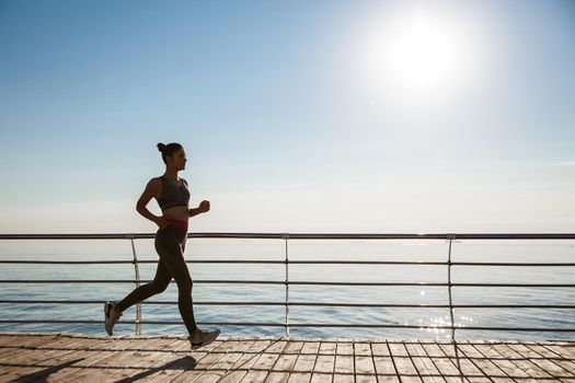 Image of attractive sportswoman jogging alone near the sea. Fitness woman running along seaside.