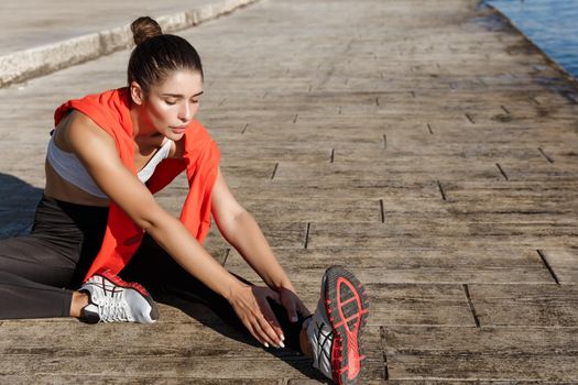 Outdoor shot of young attractive female athlete stretching leg before run, workout alone near the sea.