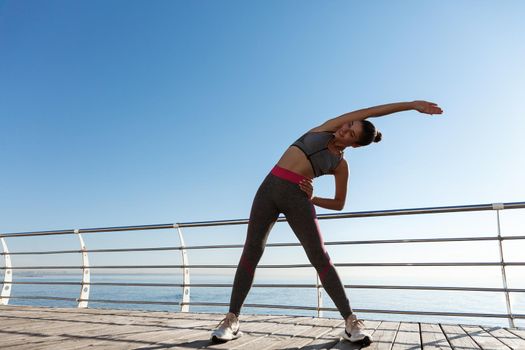Outdoor shot of attractive fitness woman training on a pier. Girl stretching before workout, standing on the seaside promenade.