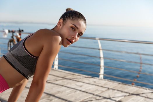 Close-up of attractive sportswoman taking a breath during workout, looking at camera and panting, jogging along seaside promenade.