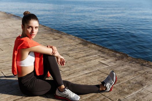 Outdoor shot of smiling fitness woman sitting near the sea and looking happy, workout on seaside promenade.