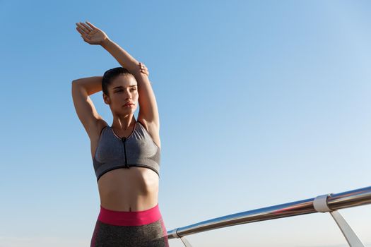Young attractive sportswoman stretching hands and warming-up before jogging on the seaside promenade. Female athlete training outdoors on a pier.