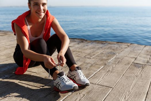 Close-up of attractive happy female athlete tying shoelaces during workout at seaside. Sportswoman training near the sea and smiling.
