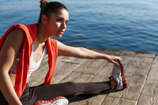 Outdoor shot of attractive fitness woman warming-up before jogging, sitting on pier and stretching legs.