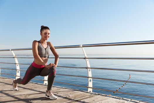 Outdoor shot of attractive and healthy fitness woman training near the sea, stretching legs before jogging.