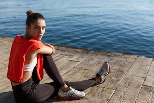 Outdoor shot of attractive sportswoman sitting near the sea and taking breath after workout, training on seaside promenade.