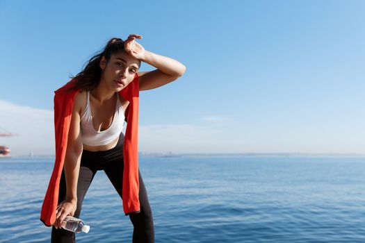 Outdoor shot of tired young sportswoman having a break near the sea, wiping sweat and panting after running.