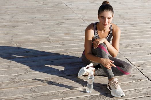 Image of attractive young female athlete having a break after workout, sitting on wooden pier and enjoying view on the sea.
