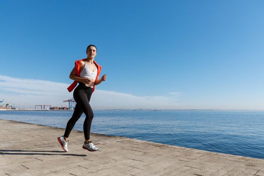 Side view of athletic young woman running along the seaside promenade. Sportswoman jogging near the sea.