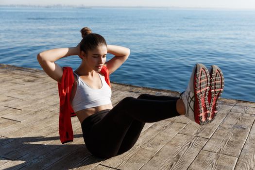 Outdoor shot of confident fitness woman working on her abs. Sportswoman doing crunches with raised legs, training near sea on a pier.