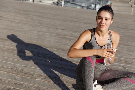 Close-up of attractive sportswoman taking a break after workout, sitting at the seaside and drinking water, looking pleased.
