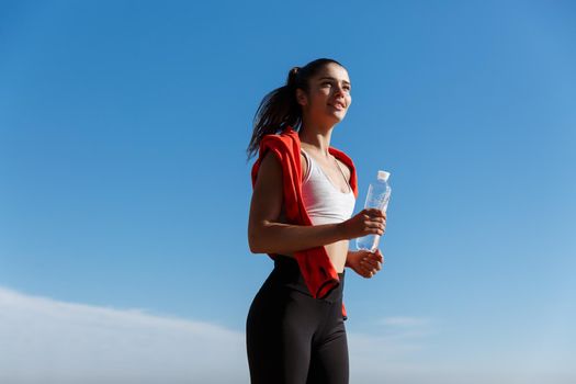 Low angle shot of happy and confident fitness woman jogging in the morning, running and smiling, holding bottle of water.