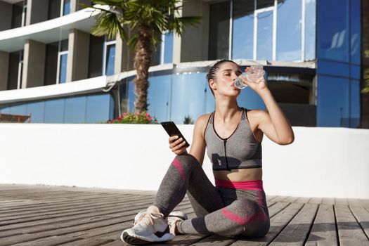 Outdoor shot of attractive fitness woman sitting on wooden pier with smartphone, having break after workout, drinking water.