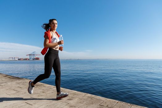 Outdoor shot of smiling sportswoman jogging and working out on seaside promenade. Fitness woman running and looking at camera.