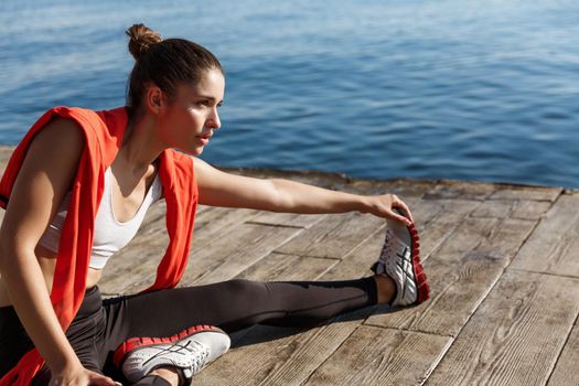 Close-up of confident sportswoman stretching legs, sitting on pier and training.
