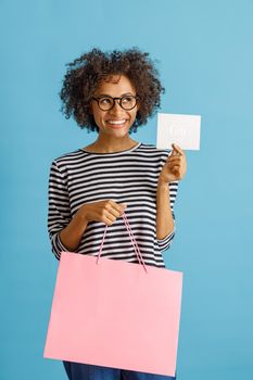 Cheerful multiracial lady looking away and smiling while holding purchases and gift certificate. Isolated on blue background