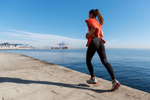 Rear view of active and healthy fitness woman running along seaside promenade. Sportswoman workout near the sea.