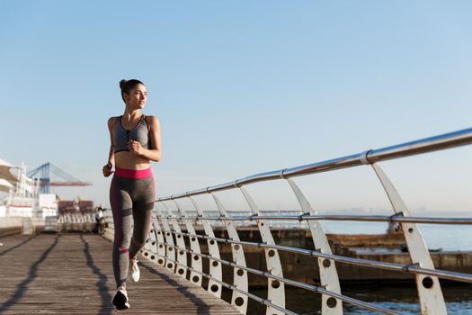 Image of attractive athletic woman in sportswear jogging in the morning, looking at sea and smiling. Female runner workout on the seaside promenade.