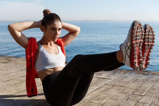 Outdoor shot of confident fitness woman working on her abs. Sportswoman doing crunches with raised legs, training near sea on a pier.