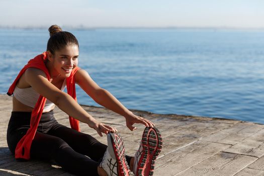 Outdoor shot of smiling confident sportswoman workout near sea. Fitness woman sitting on wooden pier and stretching legs.
