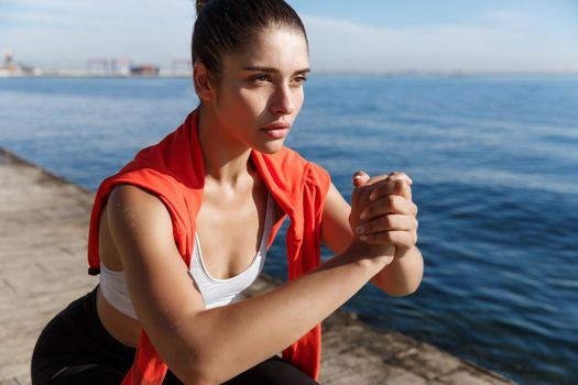 Close-up of confident and focused fitness woman looking at the sea, doing squats and workout on fresh air.