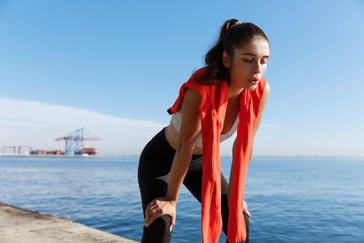 Outdoor shot of tired fitness woman panting and taking a breath after jogging, standing on a pier with sea behind her.