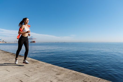 Outdoor shot of happy sportswoman jogging in the morning on a pier, running forward and smiling.
