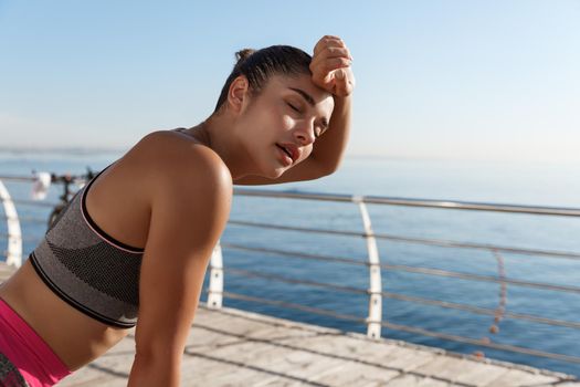 Image of beautiful young fitness woman panting and wiping sweat off forehead after jogging, finish run or workout, standing on a pier.