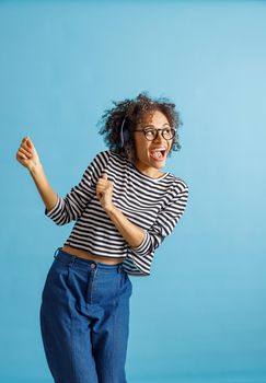 Cheerful multiracial lady in headphones enjoying favorite songs and smiling while dancing in studio. Isolated on blue background