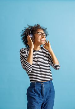 Joyful multiracial lady in headphones enjoying favorite songs and laughing. Isolated on blue background