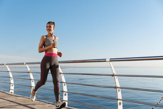 Outdoor shot of happy fitness woman running along seaside and smiling.