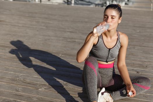 Close-up of attractive sportswoman taking a break after workout, sitting at the seaside and drinking water, looking pleased.
