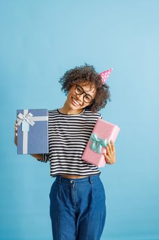 Cheerful multiracial lady in party cone hat looking at camera and smiling while holding two gift boxes. Isolated on blue background