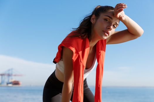 Close-up of attractive young fitness woman taking a break during jogging, panting and wiping sweat off forehead, looking tired at camera, standing near sea.