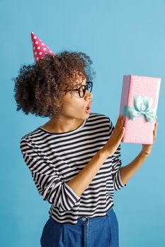 Astonished multiethnic lady in party cone hat holding gift box with ribbon. Isolated on blue background