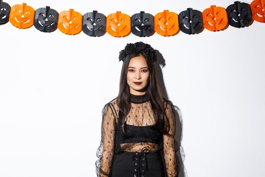 Portrait of elegant asian woman in witch costume, looking confident and standing against pumpkin streamers, decorations for halloween over white background.