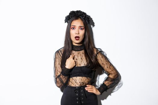Insulted and confused asian woman in halloween costume pointing at herself, wearing black dress of evil witch on party, standing over white background.