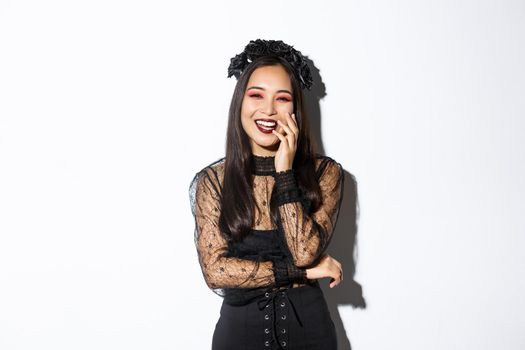 Image of happy good-looking asian woman in gothic dress and black wreath, laughing at halloween party, standing over white background.