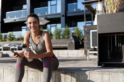 Outdoor shot of attractive fitness woman sitting with mobile phone and drinking water.