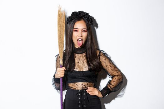Carefree sassy asian woman enjoying halloween party, wearing witch costume and holding broom, showing tongue, standing over white background.