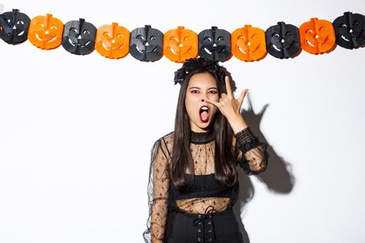 Image of funny asian woman in witch costume grimacing, wearing gothic lace dress and standing against pumpkin streamers, decorations for halloween.