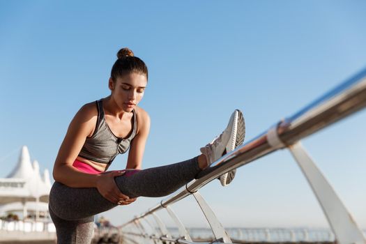 Young attractive female runner warming-up before training outdoors. Sportswoman stretching leg, prepare for jogging on the seaside promenade.