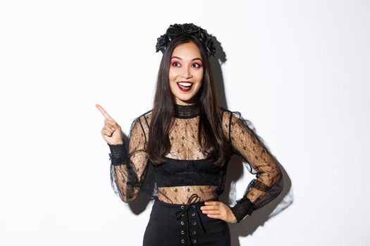 Excited happy asian woman in black lace dress and wreath looking amazed at upper left corner, pointing finger at your halloween promo banner, standing over white background.
